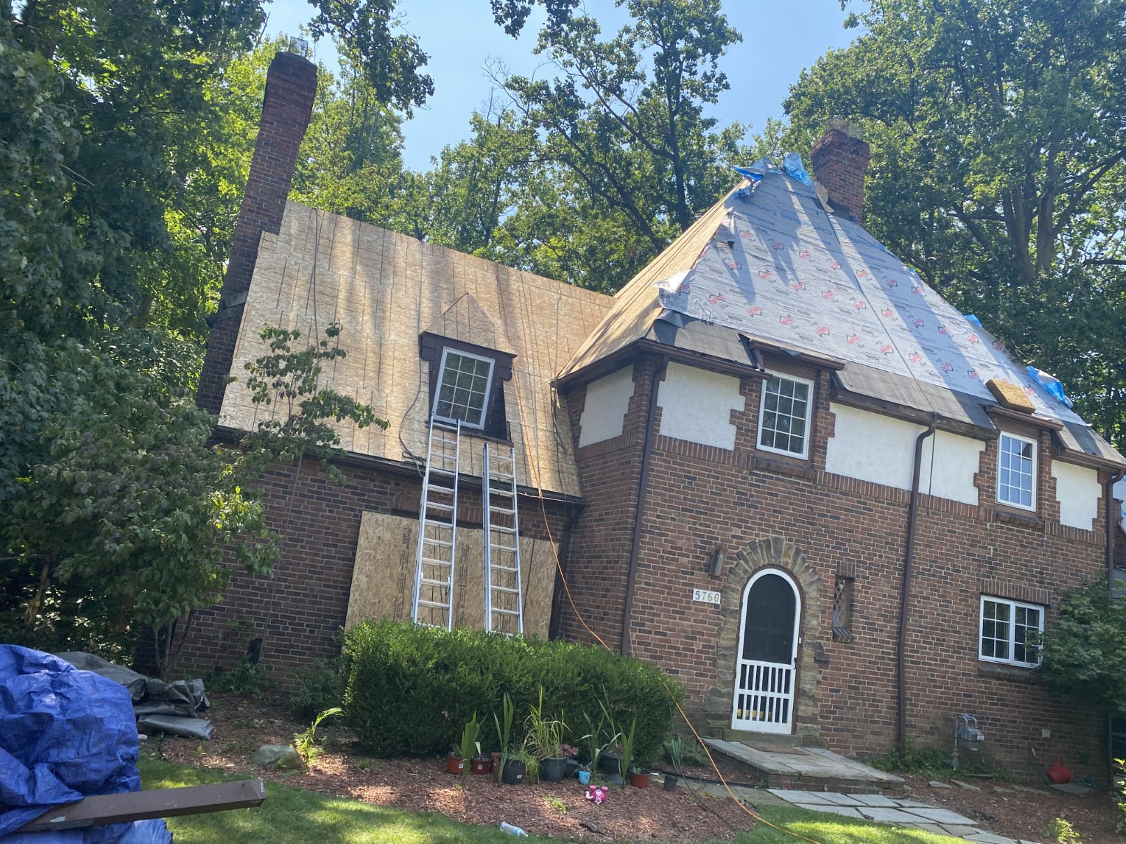 A brick Tudor home with a roof mid-project, tan underlayment and blue tarp in place