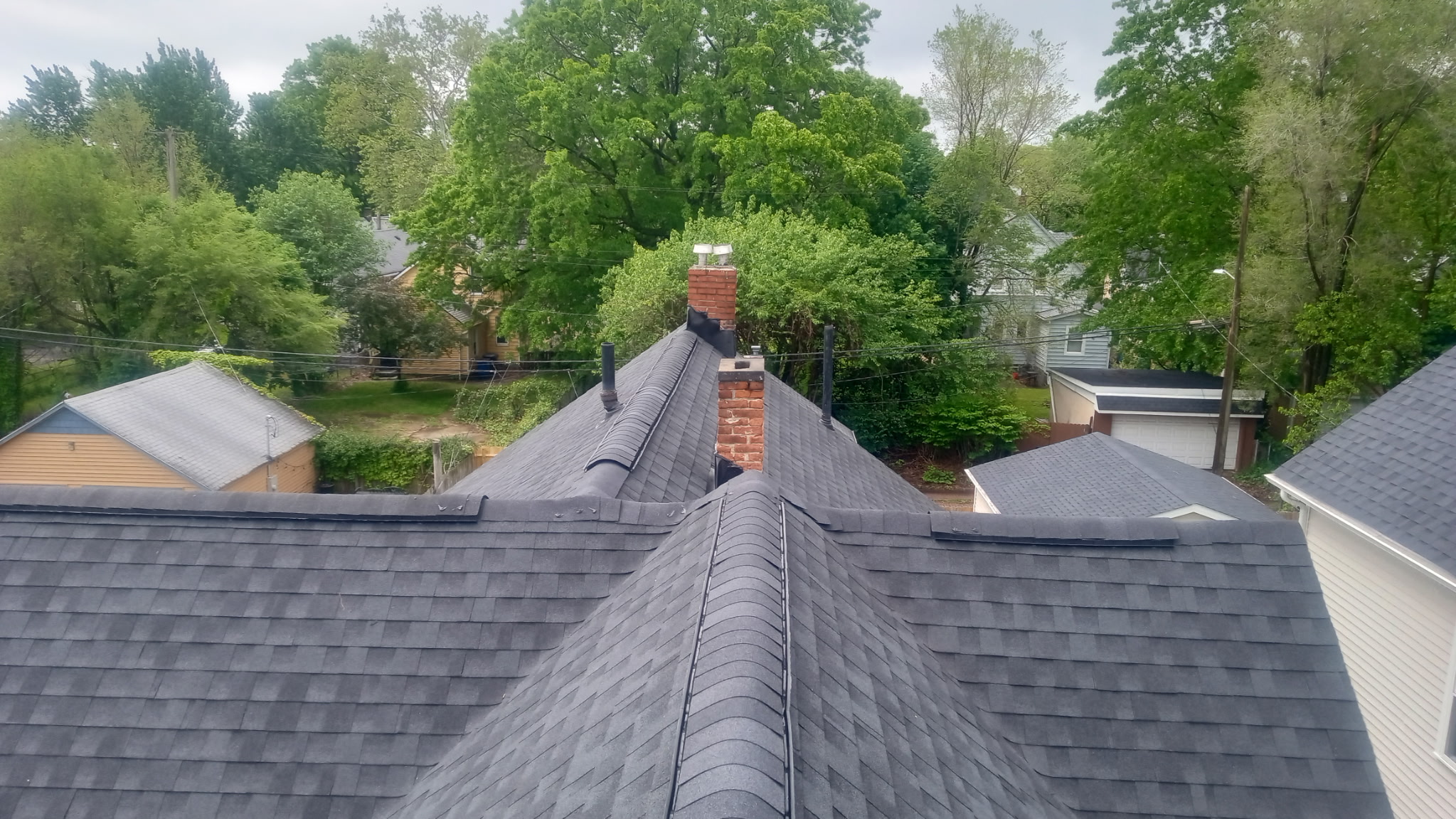 A finished grey shingle roof with a brick chimney, seen from above over the treetops